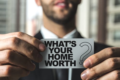 Bearded man in a suit holding a piece of paper that reads, "What's Your Home Worth?"