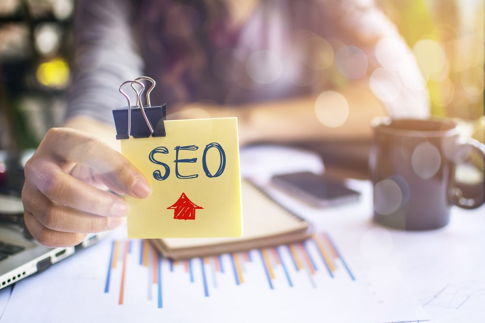 Female is sitting outside holding up a yellow post it note that is clipped and reads SEO with a red arrow pointing up