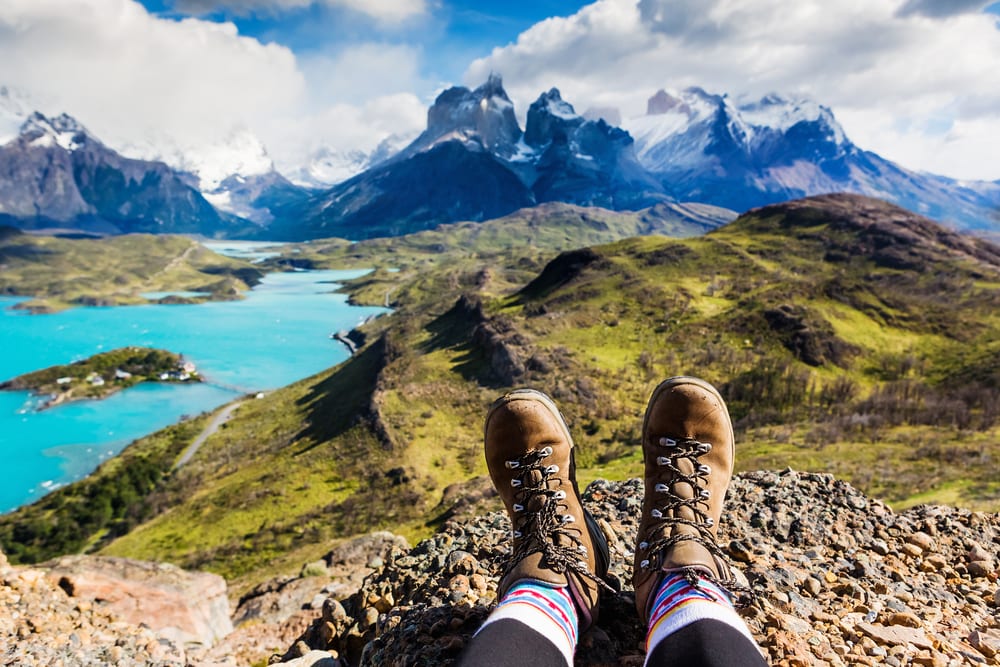 Someone is relaxing with their feet showing in hiking boots with majestic views of the mountains, water, and grassy hills