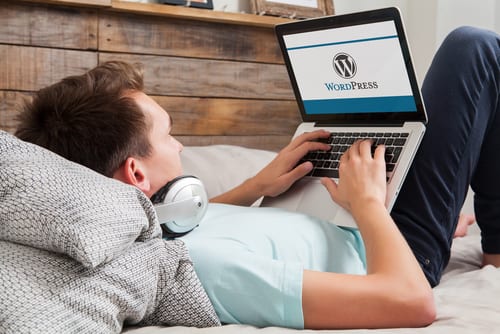 Man is laying back on pillows with headphones around the neck and has his computer propped up with wordpress on the screen
