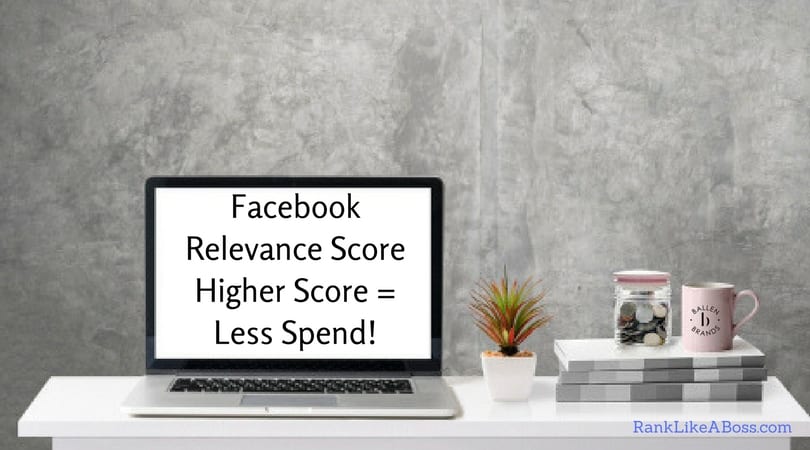 Computer is on a table with a plant, books, money jar, coffee mug. Grey wall with a lot of blank space. Computer screen reads "Facebook Relevance Score Higher Score = Less Spend!"
