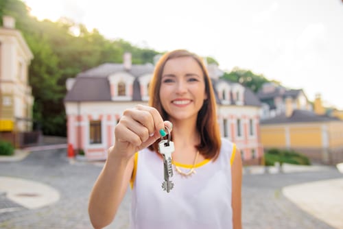 Young Real Estate Agent is holding up a pair of keys as if she sold her first house. She is standing in front of a beautiful large home