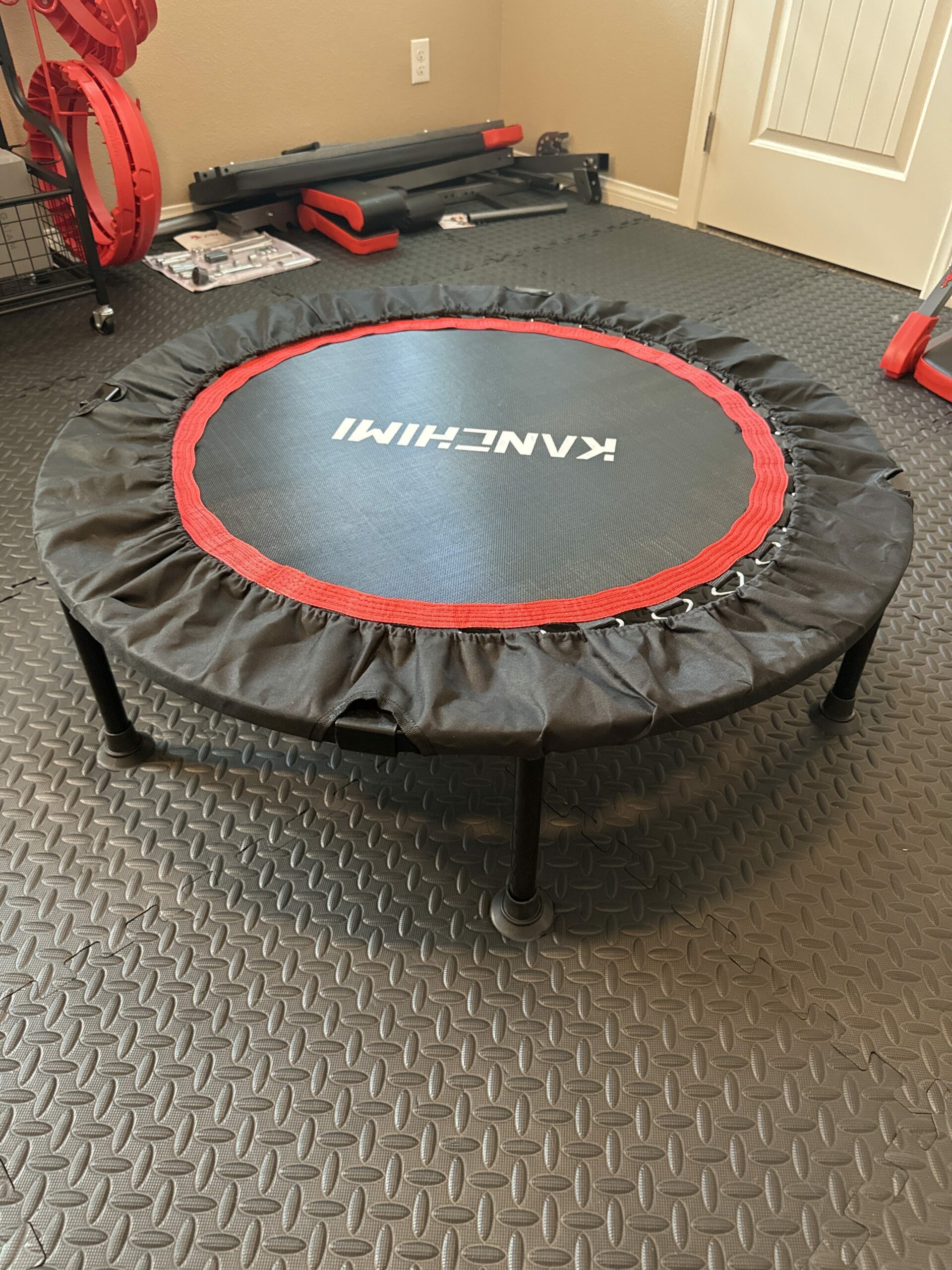 The rebounder is designed with a sturdy black mat and red trim, offering a striking contrast. It is equipped with robust legs that ensure stability during use. The surface of the rebounder is taut, providing an effective and safe bouncing experience. The compact size makes it ideal for home workouts, fitting comfortably in smaller spaces while still allowing for a wide range of exercises.