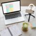 Laptop, phone on a tripod, and a content planning notebook on a marble desk — how to become a content creator